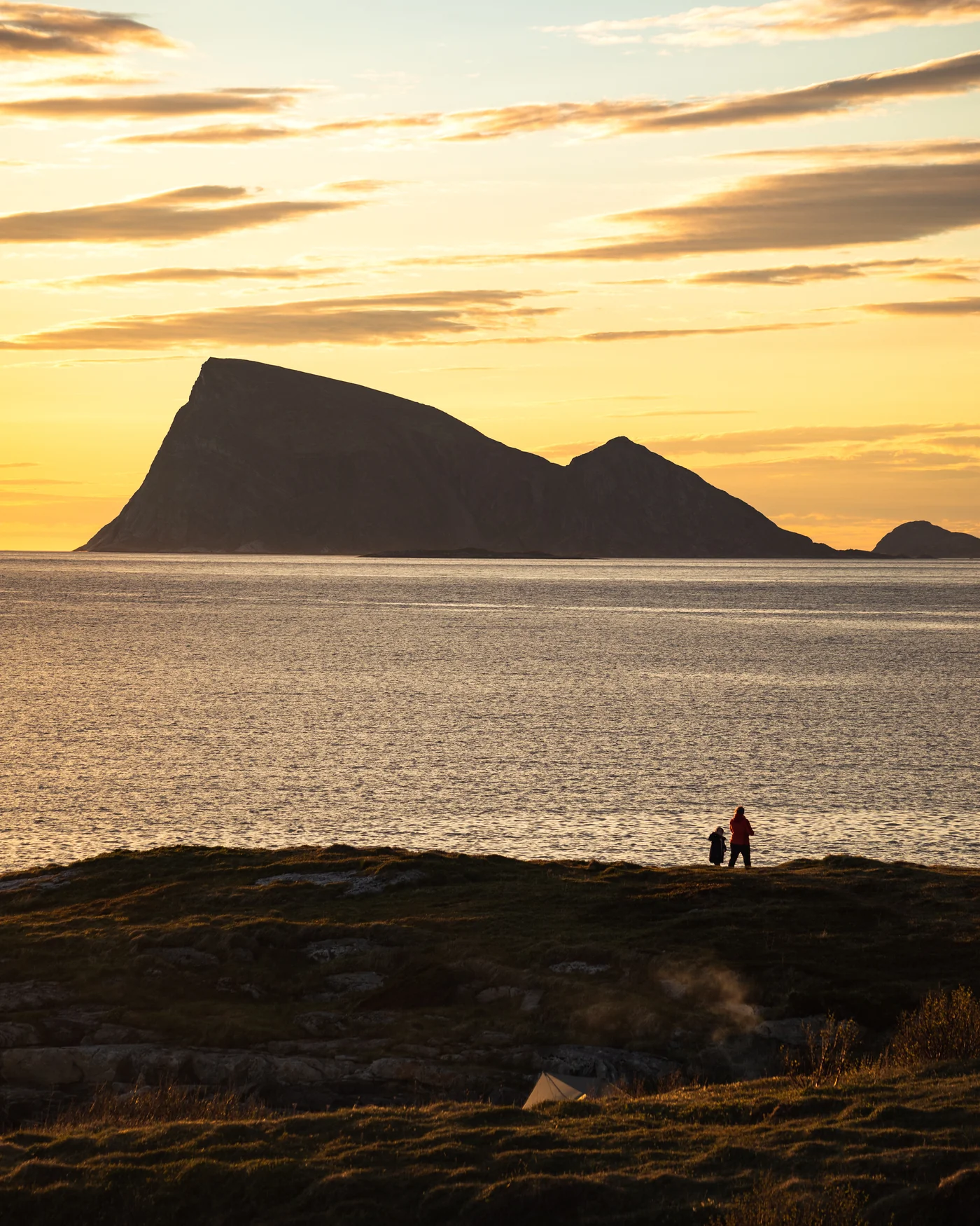Two figures on the coast outside Tromsø, dwarfed by the silhouette of Håja island under a golden midnight sky
