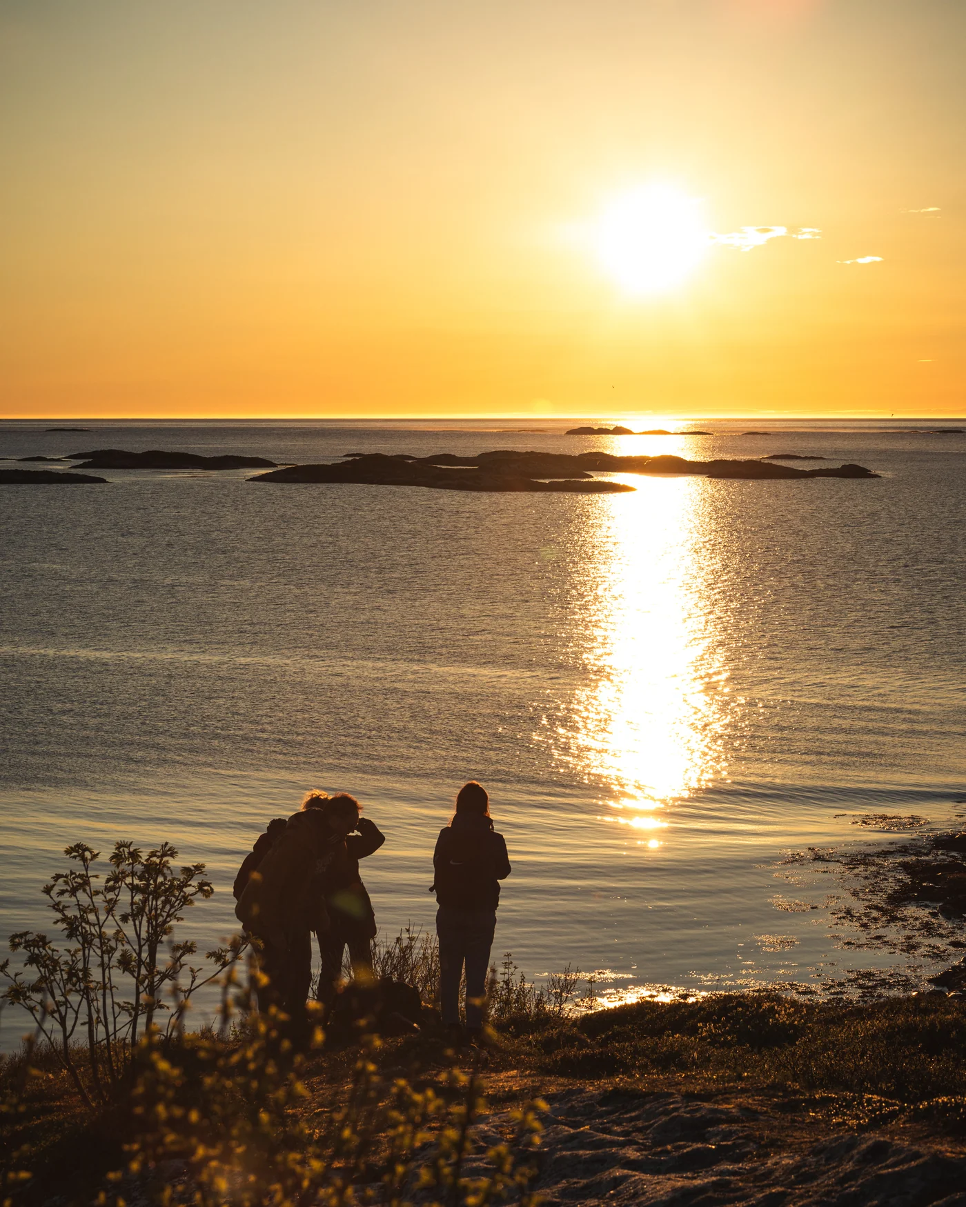 Three people standing on the coast outside Tromsø watching the midnight sun reflect across the sea