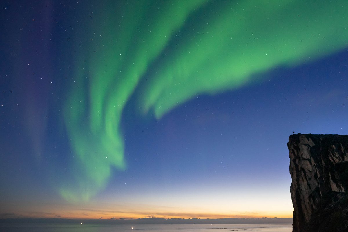 Green aurora over a sea cliff at twilight with a gold horizon near Tromsø