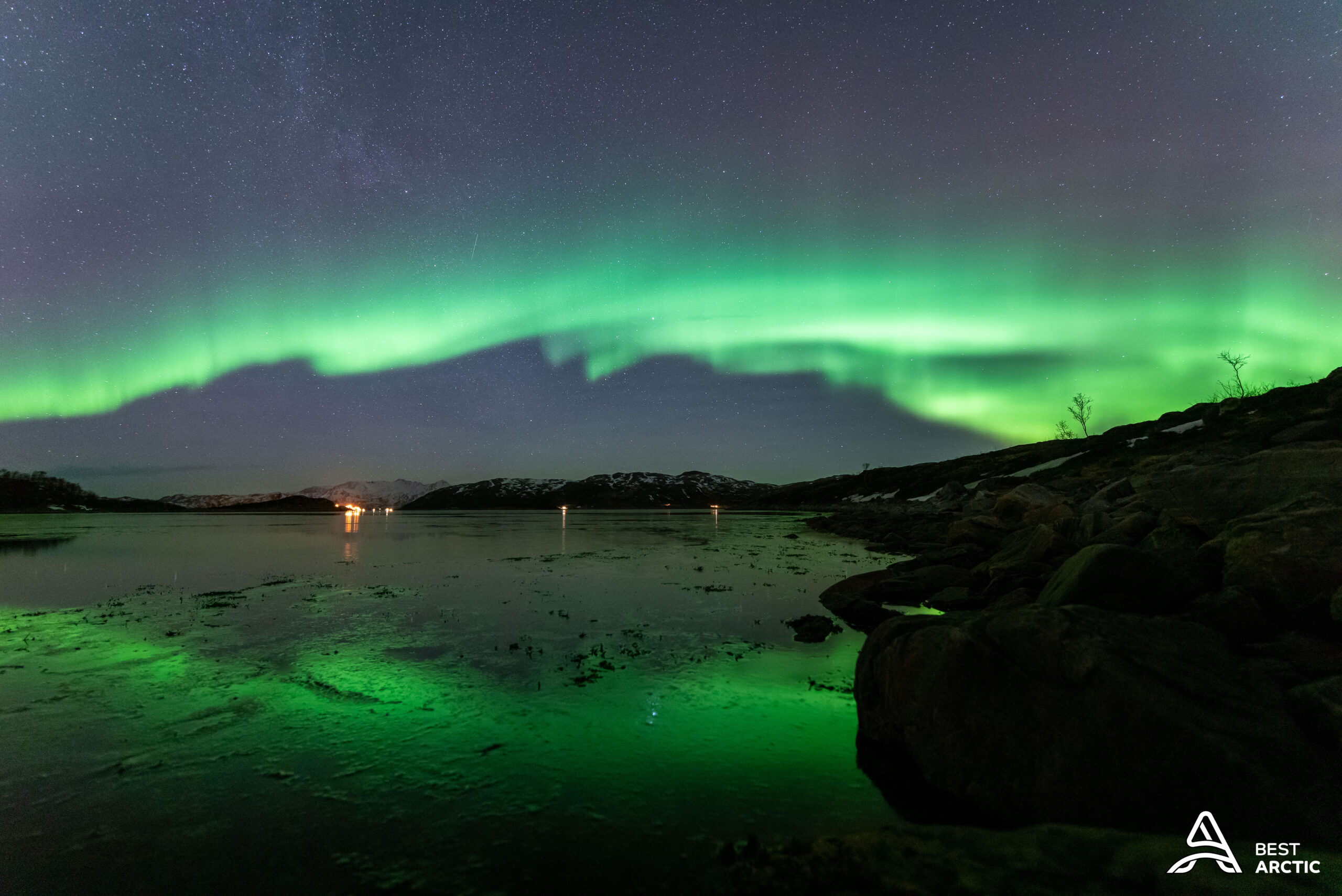 Green aurora arcing over a fjord with snow-covered mountains near Tromsø in February