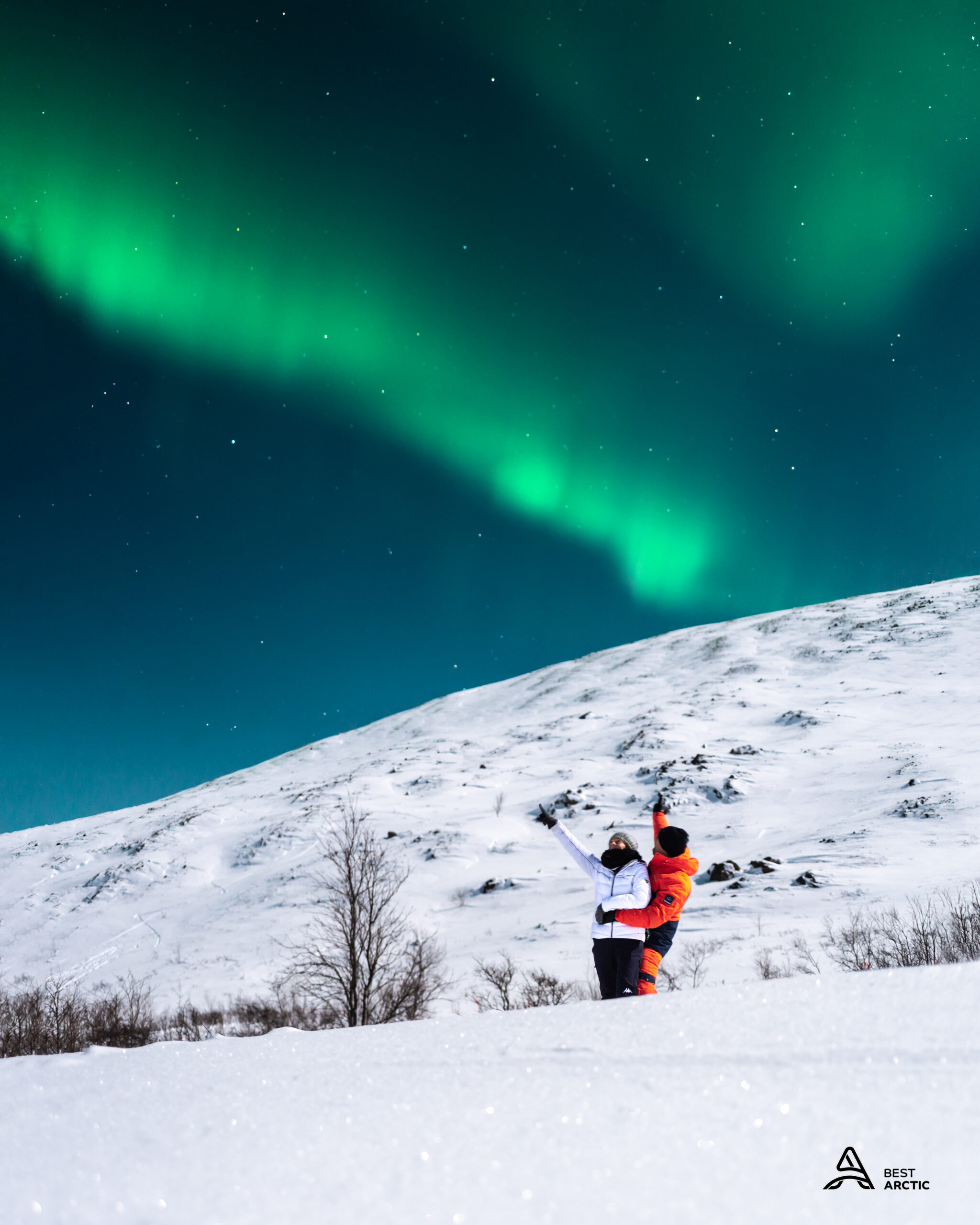 Two people in winter clothing pointing at the green aurora over snowy hills near Tromsø