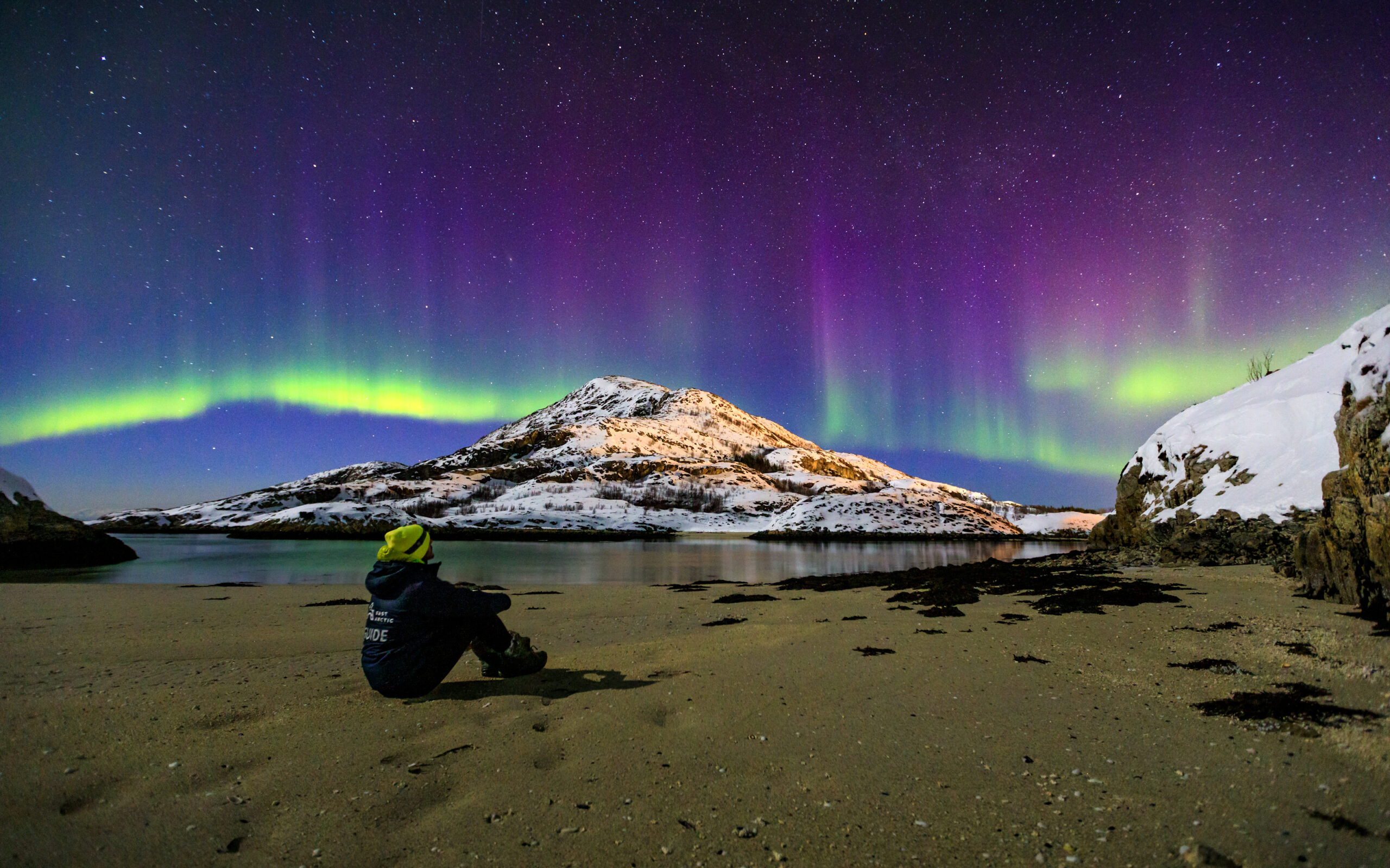 A guide watching the purple and green aurora over a fjord and snowy mountains near Tromsø
